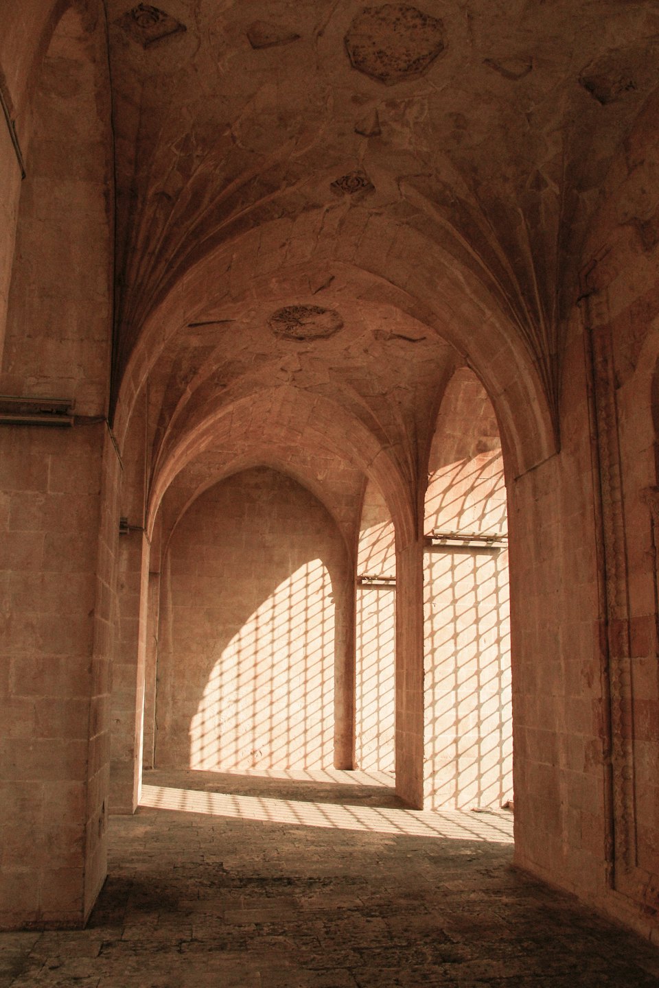 Corridoio in pietra illuminato dal sole con aperture ad arco e ombre a diamante.